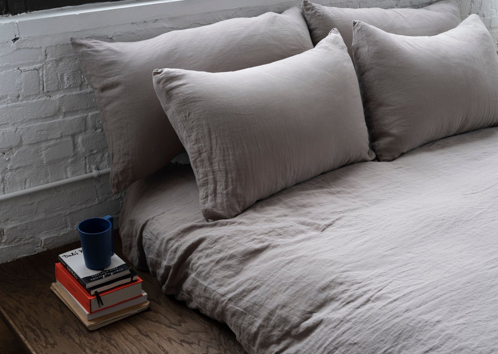 Gray bedding with pillows on a bed next to a stack of books and a blue cup.
